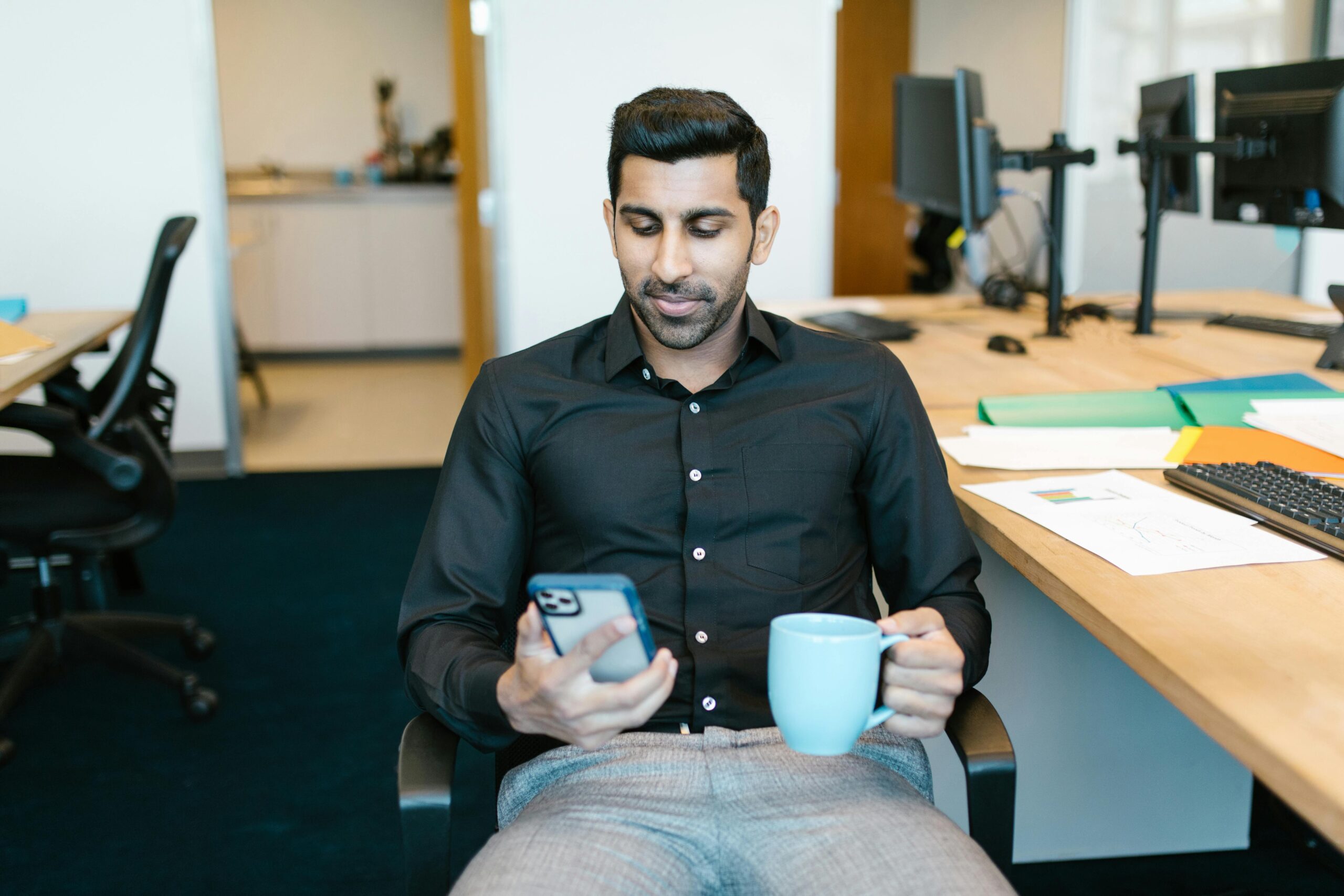 Man in an office setting using smartphone and holding coffee, dressed in black shirt.
